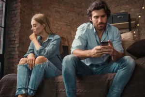 Casal brigado sentado no sofá. Mulher brava olhando para o lado e homem mexendo no celular.