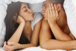 Casal apaixonado fazendo biquinho de beijo debaixo do edredon