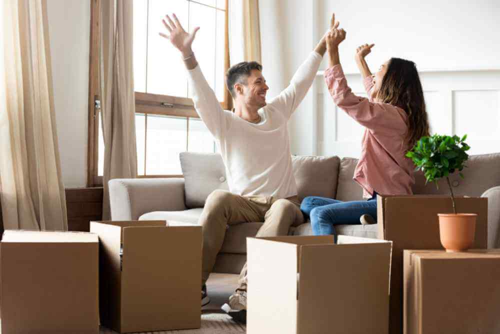Mulher comemorando algo com seu marido sentados no sofá de casa com caixas abertas no meio da sala