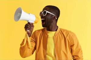 Homem negro com roupa amarela e óculos branco gritando em um megafone