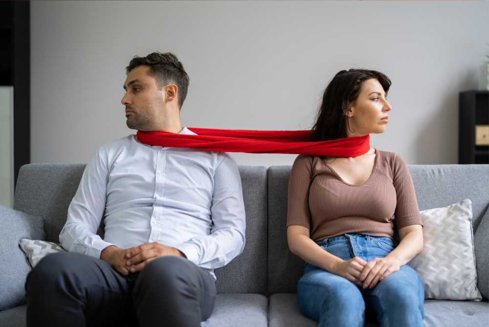 Casal afastado sentado no sofá com um pano vermelho os segurando pelo pescoço