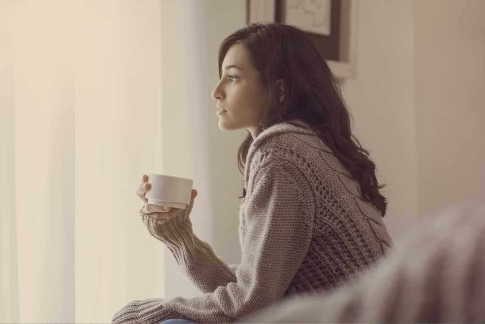 Mulher sentada no sofá tomando uma xícara de café olhando para o horizonte