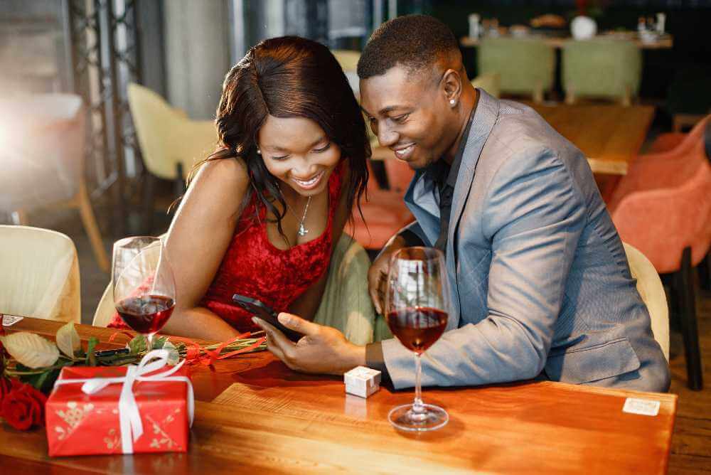 Casal em um encontro romântico no restaurante. Homem negro e mulher olhando para smartphone juntos. Mulher usando vestido elegante vermelho e homem terno azul.